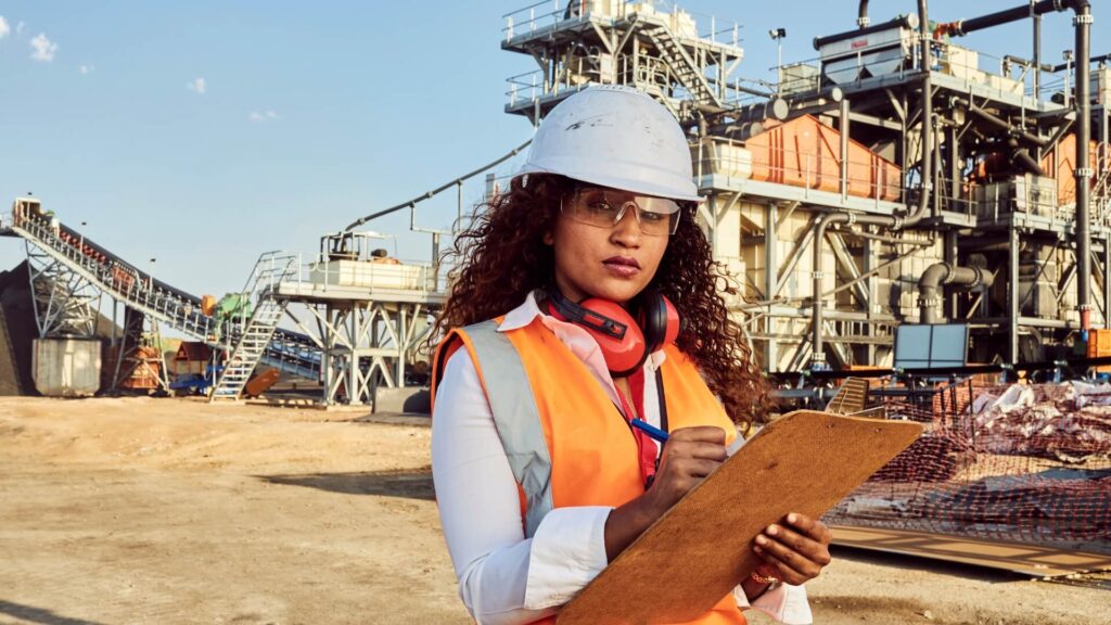 A confident female project manager wearing a white hard hat, safety glasses, and an orange safety vest stands at a construction site. She holds a clipboard and pen, appearing to be taking notes or inspecting the site. Industrial machinery and conveyor belts are visible in the background under a clear sky.