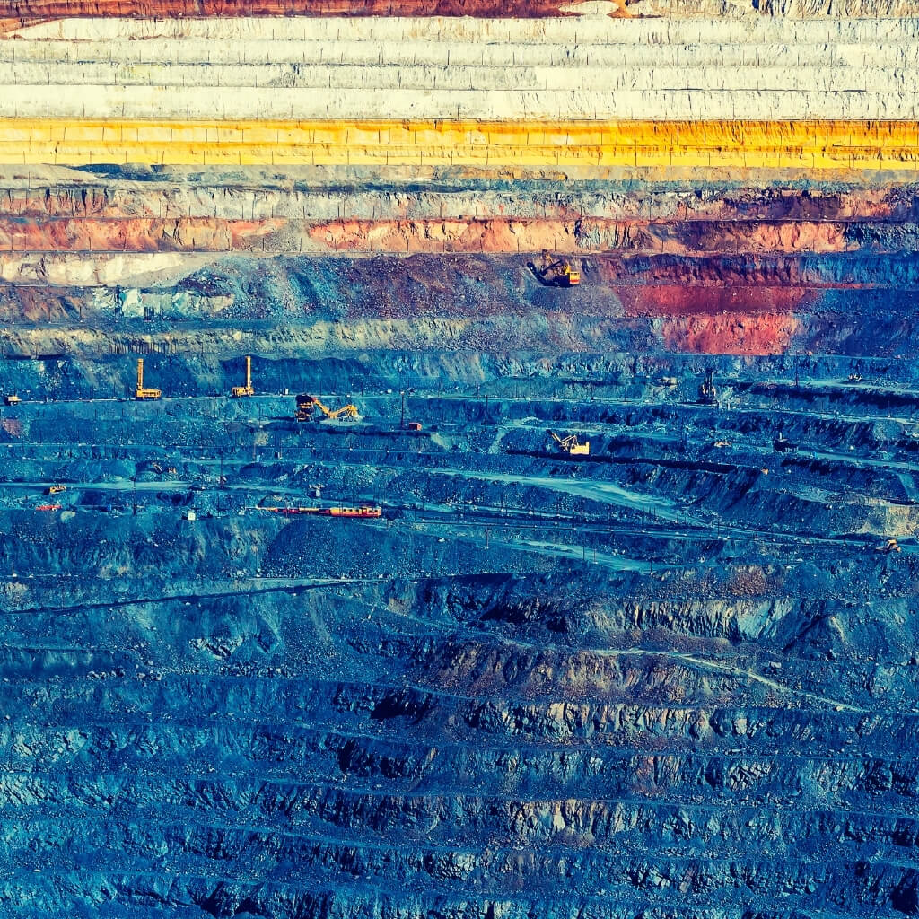 An aerial view of a large-scale mining site, revealing the striking layers of rock and earth in an open-pit mine. The walls show a gradient of colors from rust and yellow to a deep blue, with small excavators at work on the different tiers. This image is an excellent representation of a massive resource extraction project.