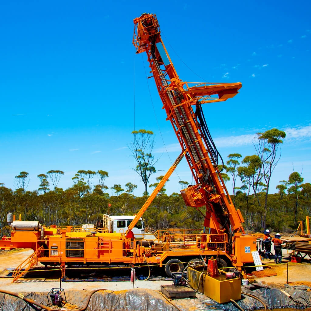 A large, bright orange drilling rig stands prominently on a dirt lot, with a few workers visible near its base. The tall rig is actively drilling, with its mast extended toward a clear blue sky. In the background, there are sparse trees and a flat landscape. The image represents geological exploration and drilling services in the mining industry.
