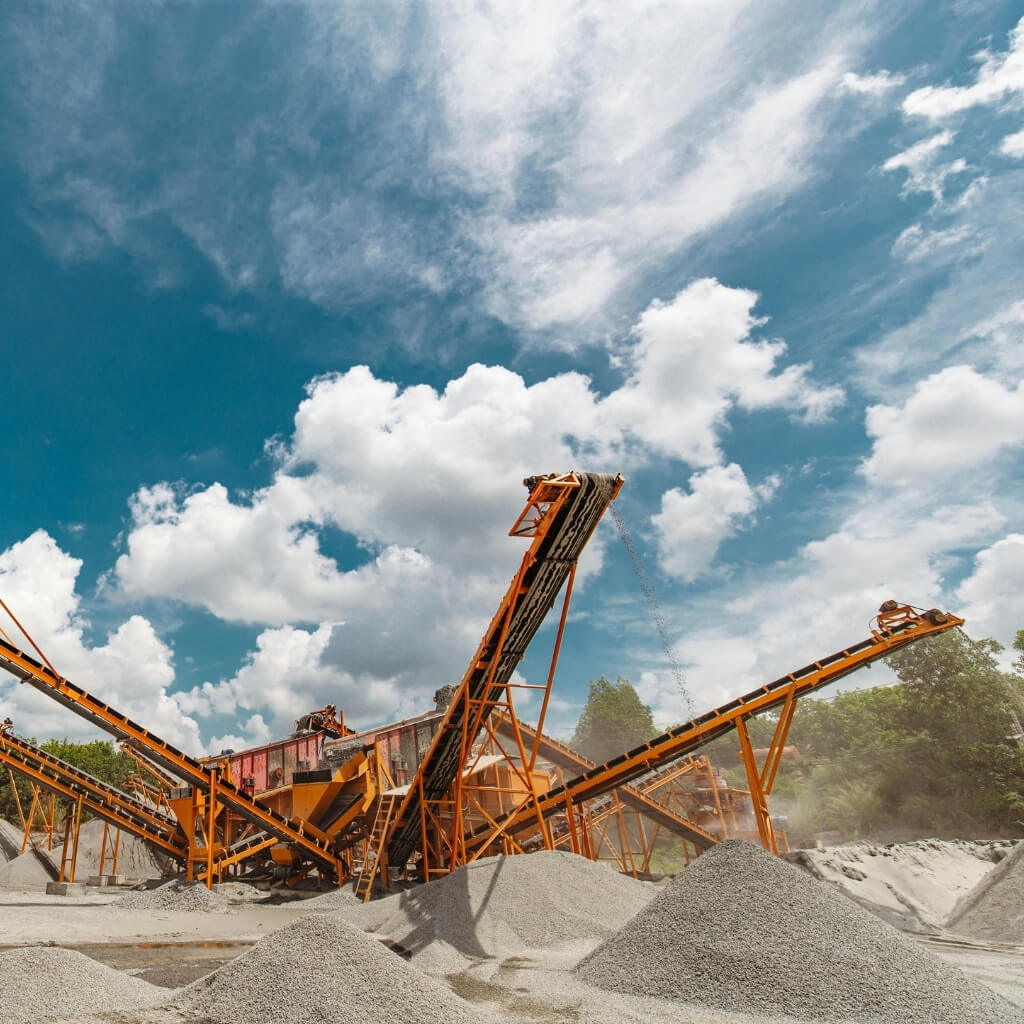 A low-angle shot of a large mineral processing plant, with several orange conveyor belts and crushing machinery forming a complex industrial structure. The equipment is surrounded by large piles of crushed rock and gravel, indicating a high volume of material processing. The scene is set under a bright blue sky with large, puffy white clouds, suggesting a sunny day. This image represents the core services of metallurgy and mineral processing.