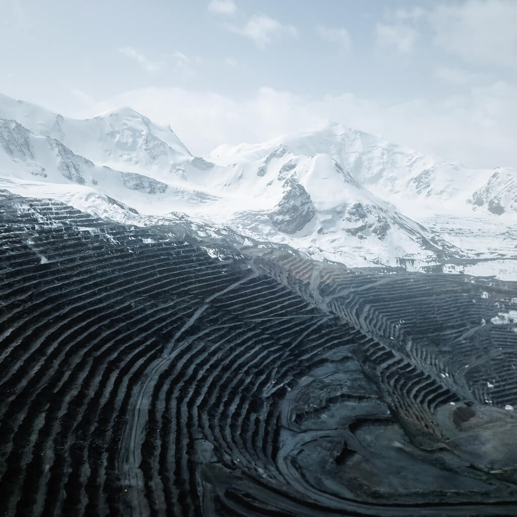An aerial view of a massive, dark open-pit mine with a terraced, circular structure that contrasts sharply with the towering, snow-covered mountains in the background. The scene is set under a cold, overcast sky, highlighting the scale and challenging environment of large-scale mining operations in remote, mountainous regions.