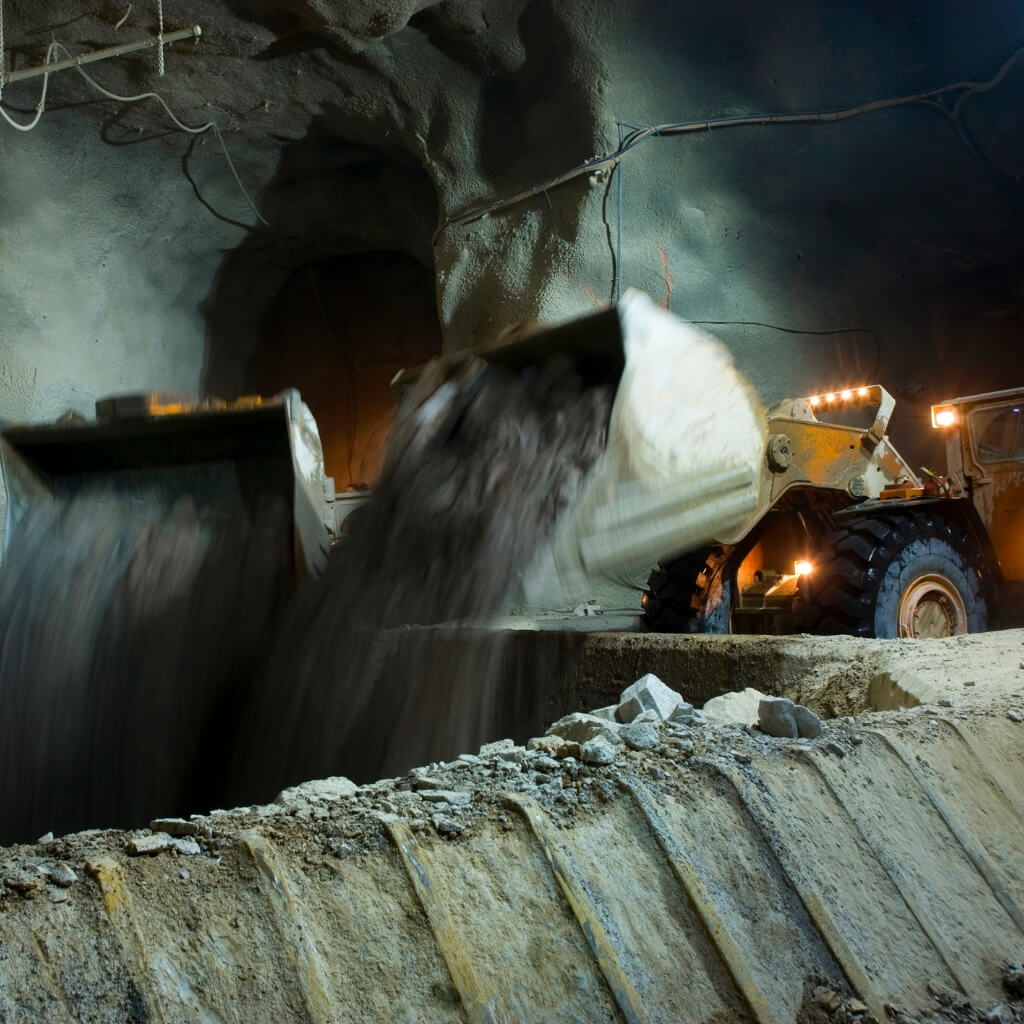 A wide-angle shot from a low perspective inside a dark underground mine tunnel. Two large scoops of earthmoving machinery, likely loaders, are visible. The one in the foreground is tipping its load of rocky material, which is in motion and blurred. The machinery is illuminated by its own lights, highlighting the dusty and rocky environment. This image represents project management and operational support in underground mining.