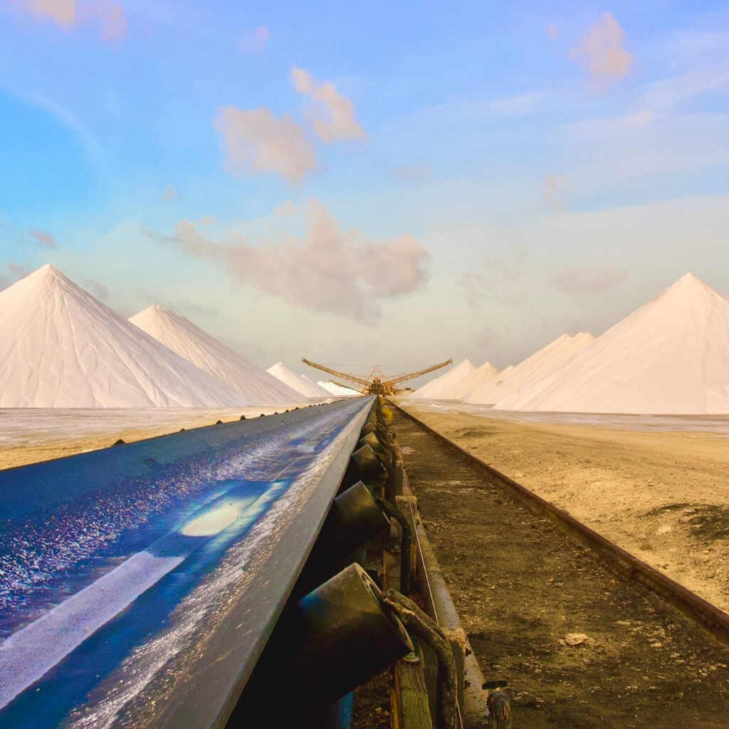 Low-angle view of a large industrial conveyor belt leading into a field of massive, white, conical salt or mineral stockpiles against a bright blue and orange sky. The scene highlights material handling and industrial automation in resource processing.